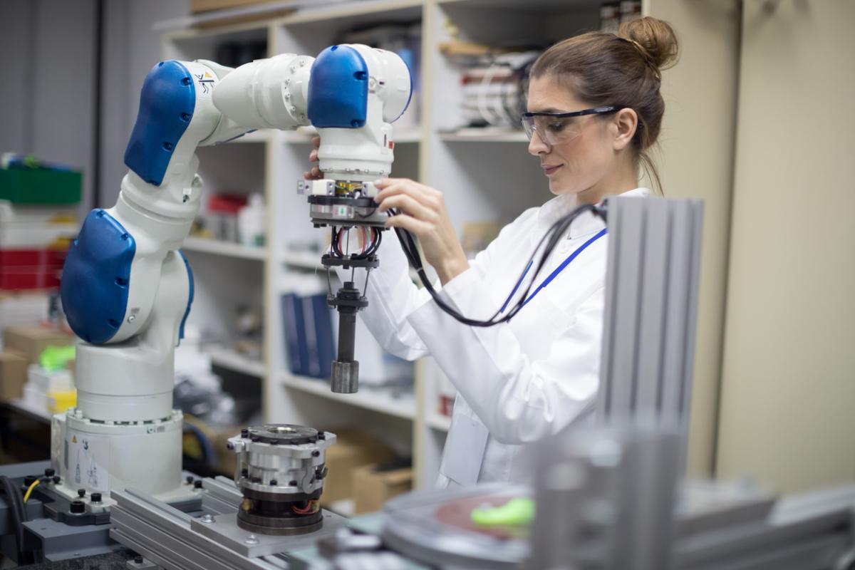 A female engineer in a white coat and safety glasses adjusts a collaborative robot.